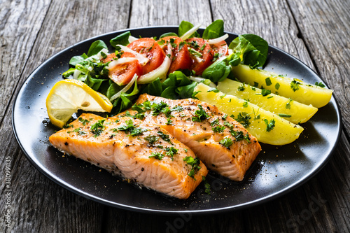 Fried salmon fillet with boiled potatoes, lemon and vegetable salad served on black plate on wooden table
