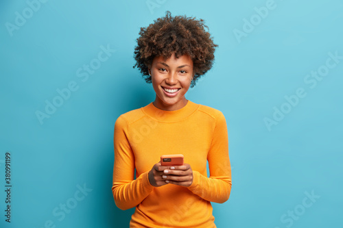 Photo of cheerful dark skinned young woman holds modern device in hands browses her favorite web page surfs social media sends sms smiles positively dressed casually isolated on blue background
