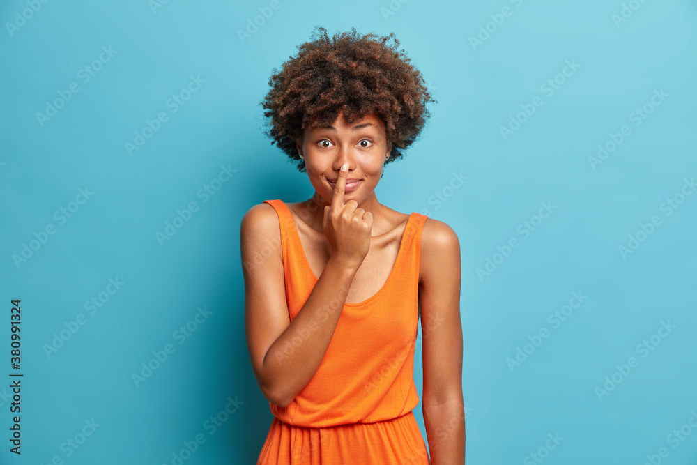 Funny dark skinned woman touches her nose and looks with happy expression dressed in orange dress poses against blue background. Positive beautiful Afro American girl makes fun smiles happily Funny dark skinned woman touches her nose and looks with happy expression dressed in orange dress poses against blue background. Positive beautiful Afro American girl makes fun smiles happily