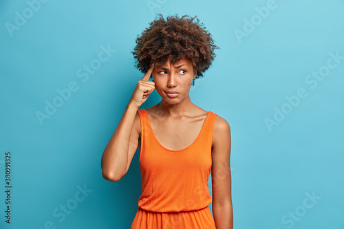 Portrait of troubled displeased beautiful young Afro American woman holds finger on temple tries to create plan in mind focus and solve puzzle concentrated on something dressed in orange dress photo