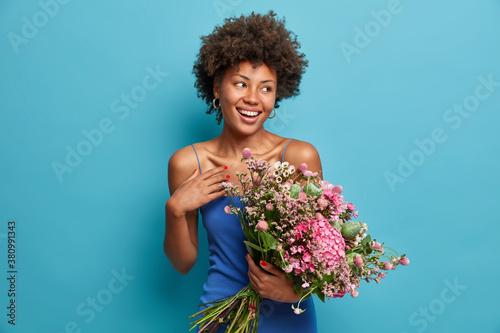 Charming romantic beautiful African American woman holds large bouquet receives flowers from secret admirer enjoys unexpected birthday surprise dressed in fashionable dress isolated on blue wall