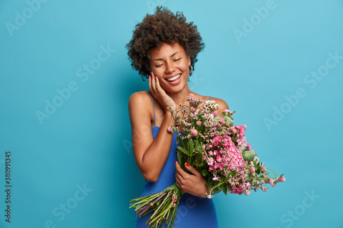 Cheerful positive dark skinned young woman smiles happily with closed eyes holds big bouquet of flowers happy to receive birthday compliment poses against blue background. 8 March celebration