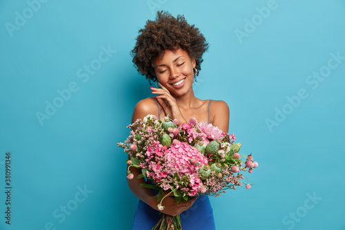 Charming tender woman stands with closed eyes and touches face gently enjoys boyfriend birthday gift surprise holds beautiful flowers isolated on blue background. Romantic dreamy lady with bouquet