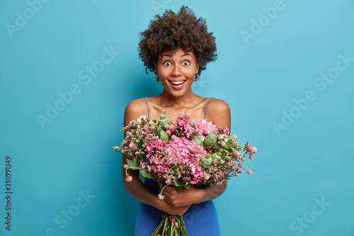Positive cheerful adorable Afro American woman smiles broadly and holds big delivery bouquet of nice flowers celebrates birthday isolated on blue background. Women holiday event celebration concept