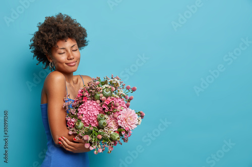 Sideways shot of romantic beautiful lady enjoys large bouquet appreciates gift receieved from boyfriend celebrates special occasion hears pleasant smell stands with closed eyes against blue wall