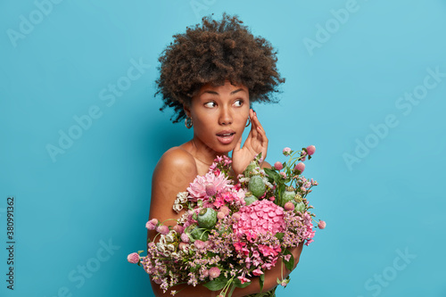 Pretty young lady has Afro hair holds big beautiful bouquet of flowers and looks with wonder away poses bare shoulders against blue background. Natural beauty celebration and feminity concept