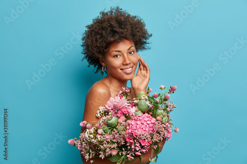 Profile side of dreamy charming woman with bare shoulders touches face gently smiles pleasantly enjoys 8 march celebration holds bouquet of flowers stands against blue background receives congrats