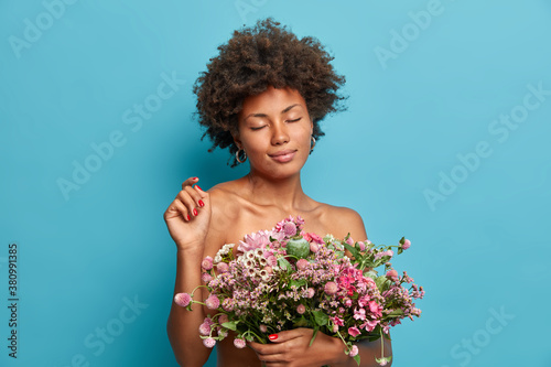 Horizontal shot of natural beautiful woman with curly hair stands indoor has romantic mood holds nice bouquet of flowers enjoys spring celebration got present from husband on wedding anniversary