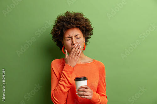 Photo of beautiful tired woman yawns and being sleepy after awakening early in morning holds disposable cup of coffee dressed in casual jumper isolated on vivid green background. Exhausted girl