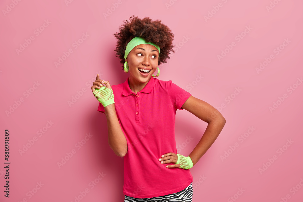 Photo of active positive Afro American woman raises hand in sport gloves and smiles happily dressed in active wear poses against pink background being always fit and healthy. Sporty lifestyle concept