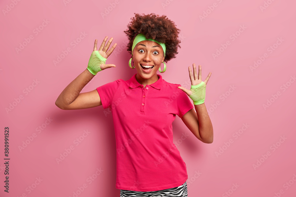 Horizontal shot of positive Afro American girl raises palms and has fun smiles broadly looks happily at camera wears headband sport gloves and t shirt isolated on pink wall leads active lifestyle Horizontal shot of positive Afro American girl raises palms and has fun smiles broadly looks happily at camera wears headband sport gloves and t shirt isolated on pink wall leads active lifestyle