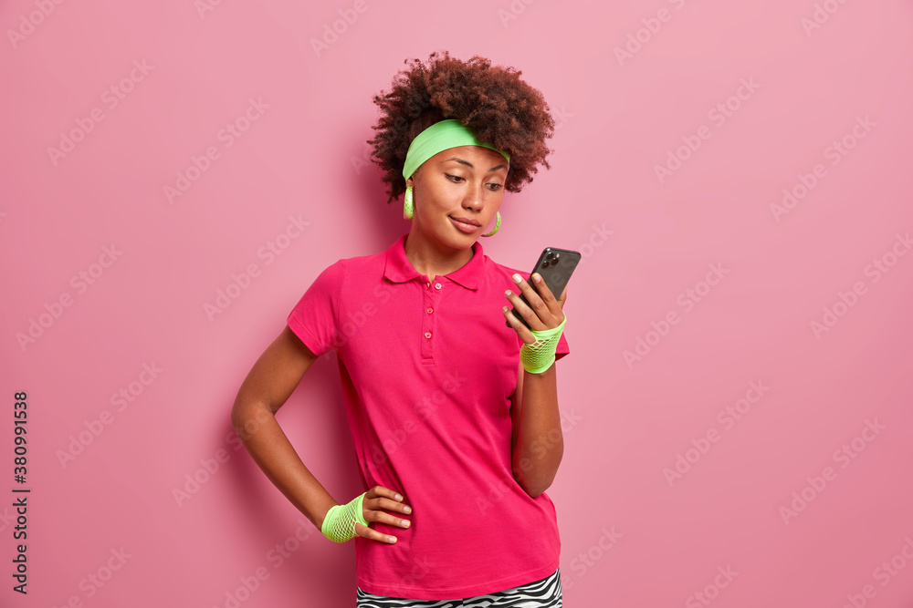 Beautiful curly haired young woman sends message on smartphone blogs in social networks uses wireless internet wears sport clothes takes break after fitness training isolated on pink background Beautiful curly haired young woman sends message on smartphone blogs in social networks uses wireless internet wears sport clothes takes break after fitness training isolated on pink background