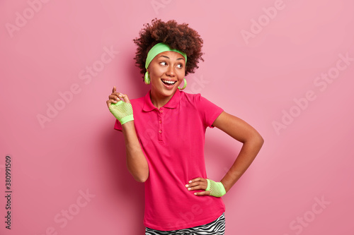 Photo of active positive Afro American woman raises hand in sport gloves and smiles happily dressed in active wear poses against pink background being always fit and healthy. Sporty lifestyle concept