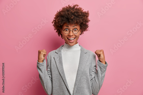 Optimistic excited businesswoman raises clenched fists and celebrates triumph or successful project fulfillment dressed in formal grey clothes wears round spectacles isolated on pink studio wall