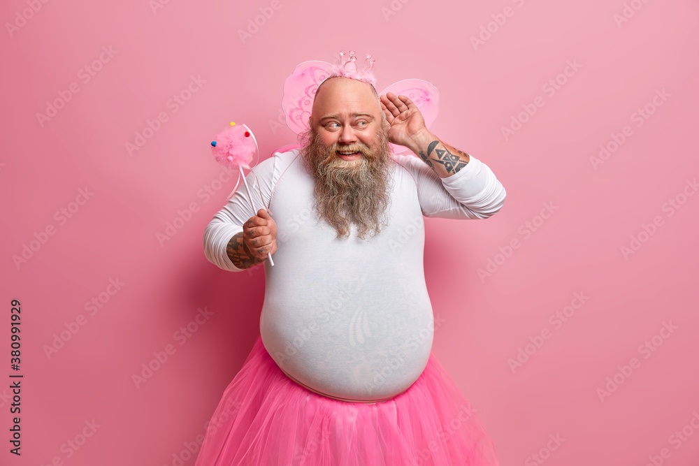 Fototapeta premium Funny overweight bearded man dressed in fairy costume to entertain guests of party keeps hand near ear and tries to overhear something holds magic wand has big belly poses against pink background