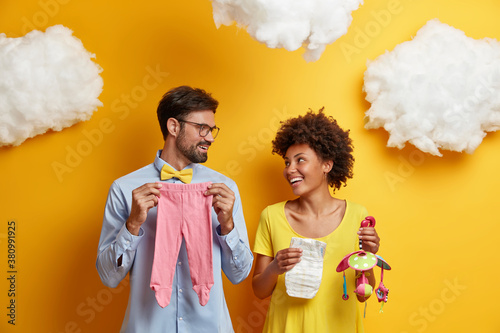 Happy couple future parents prepare for parenthood and giving birth to baby pose with infant items looks cheerfully at each other pose together against yellow wall. Healthy pregnancy family life