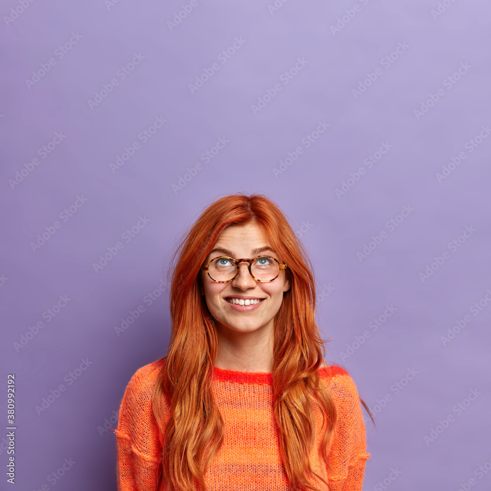 Pleased curious redhead European woman concentrated above with broad smile wears optical glasses dressed casually isolated on purple background copy space for your advertisement or promotion Pleased curious redhead European woman concentrated above with broad smile wears optical glasses dressed casually isolated on purple background copy space for your advertisement or promotion