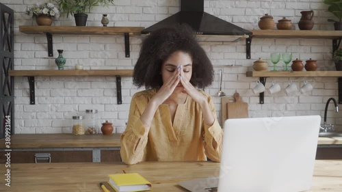 Sad depressed young mixed race female sitting at kitchen table with papers and laptop, having tired worried look, massaging eyes while calculating family budget, stressed with financial problems