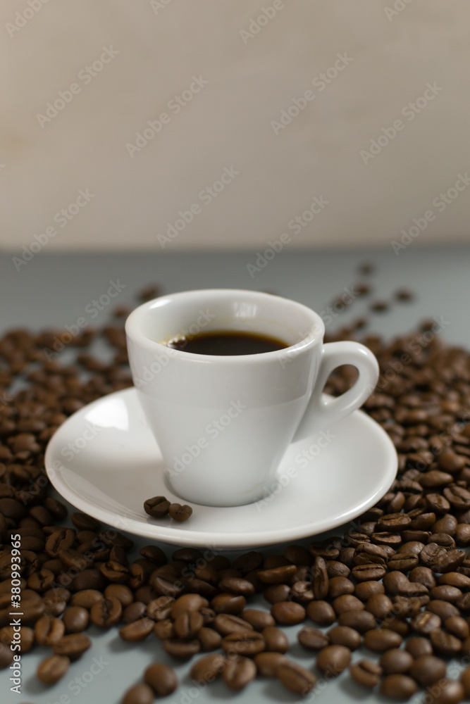 Fototapeta premium White cup with espresso on a white saucer among coffee beans on a gray background. Copy space. Selective focus. Vertical orientation.