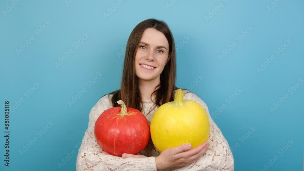 Cheerful positive young woman embraces two ripe pumpkins, smiles pleasantly, dressed in white sweater, isolated on blue studio background. Autumn harvesting, food and Halloween holiday concept. Cheerful positive young woman embraces two ripe pumpkins, smiles pleasantly, dressed in white sweater, isolated on blue studio background. Autumn harvesting, food and Halloween holiday concept.