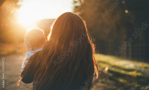 Blurred mom with child stands on the mountain at sunset. Orange rays from the sun provide backlighting and bokeh. Mother and baby concept, inspiration. Close-up. Back view
