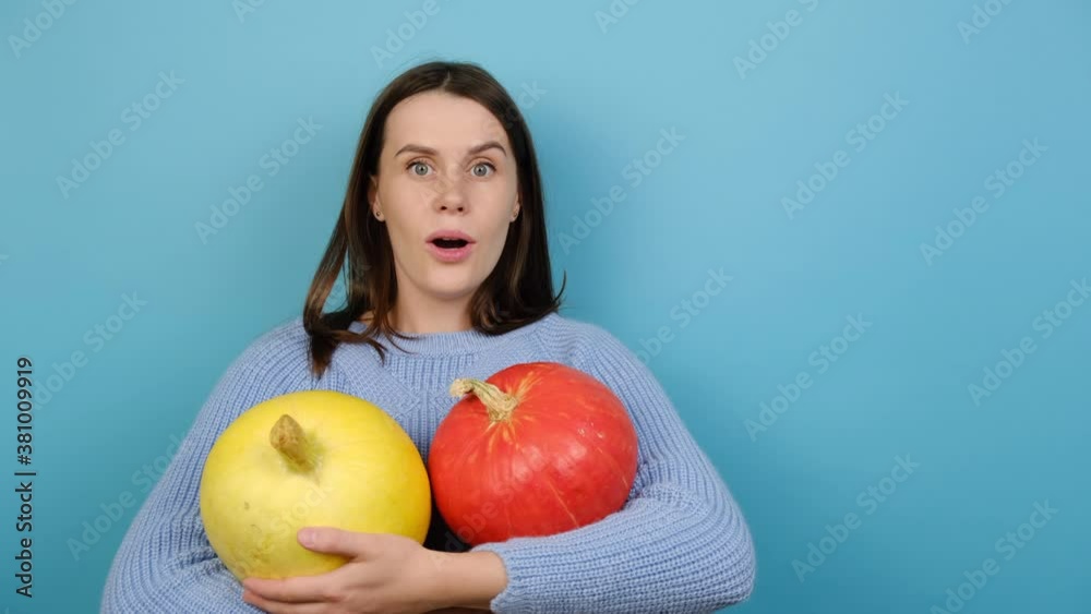 Shocked young woman holds small pumpkins, wears white knitted sweater, looks aside, keeps mouth open, isolated on blue background with copy space for advertisement. Halloween and autumn concept Shocked young woman holds small pumpkins, wears white knitted sweater, looks aside, keeps mouth open, isolated on blue background with copy space for advertisement. Halloween and autumn concept