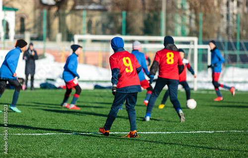 Boys in red sportswear running on soccer field with snow on background. Young footballers dribble and kick football ball in game. Training, active lifestyle, sport, children winter activity