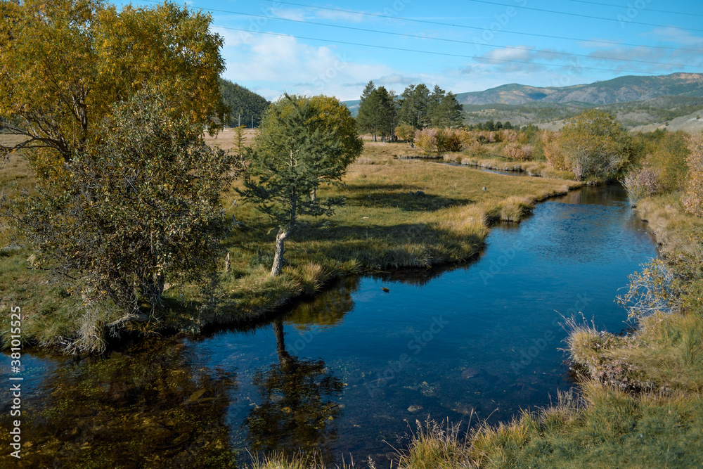 Bright blue winding river flows in grass, bushes, trees among the mountains and blue sky with clouds. Reflections in the water. Autumn landscape