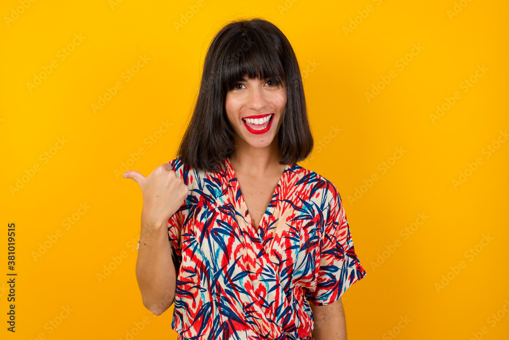 Omg concept. Stupefied caucasian female with surprised expression, opens eyes and mouth widely, points aside with thumb, shows something strange on gray background. Advertisement concept. Omg concept. Stupefied caucasian female with surprised expression, opens eyes and mouth widely, points aside with thumb, shows something strange on gray background. Advertisement concept.