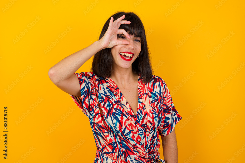 Beautiful young blonde woman with happy face smiling doing ok sign with hand on eye looking through finger. Wearing casual clothes and standing. Beautiful young blonde woman with happy face smiling doing ok sign with hand on eye looking through finger. Wearing casual clothes and standing.