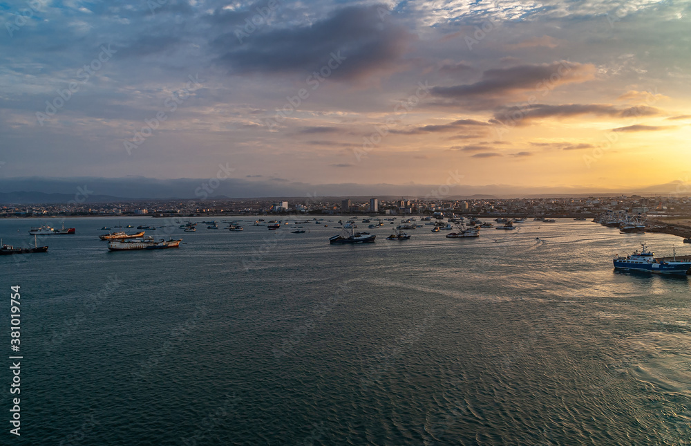 Fototapeta premium Manta, Ecuador - December 2, 2008: Sunset over harbor shows dark ocean water up front with many small boats on top, light over cityscape as central belt. Cloudscape and mountain range in back.