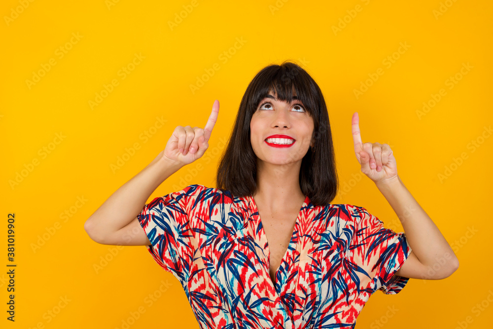 Horizontal shot of successful friendly looking young dark haired woman with bright make up exclaiming excitedly, pointing both index fingers up, indicating something shocking on blank copy space wall. Horizontal shot of successful friendly looking young dark haired woman with bright make up exclaiming excitedly, pointing both index fingers up, indicating something shocking on blank copy space wall.