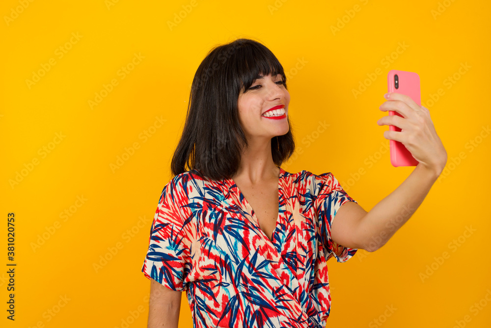 Isolated shot of pleased cheerful young woman with dark hair, makes selfie with mobile phone, dressed in casual sweater, isolated over beige background. People, technology and leisure concept Isolated shot of pleased cheerful young woman with dark hair, makes selfie with mobile phone, dressed in casual sweater, isolated over beige background. People, technology and leisure concept