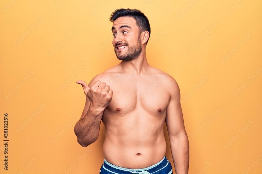 Young handsome man with beard wearing sleeveless t-shirt standing over yellow background smiling with happy face looking and pointing to the side with thumb up.