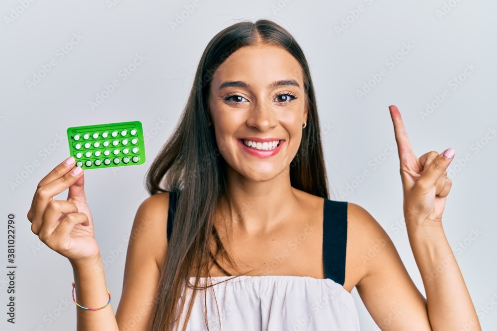 Young hispanic woman holding birth control pills smiling with an idea or question pointing finger with happy face, number one Young hispanic woman holding birth control pills smiling with an idea or question pointing finger with happy face, number one