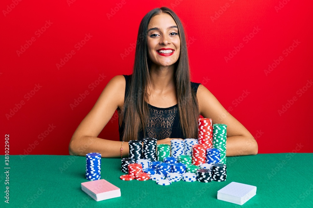Young hispanic woman sitting on the table playing poker looking positive and happy standing and smiling with a confident smile showing teeth Young hispanic woman sitting on the table playing poker looking positive and happy standing and smiling with a confident smile showing teeth