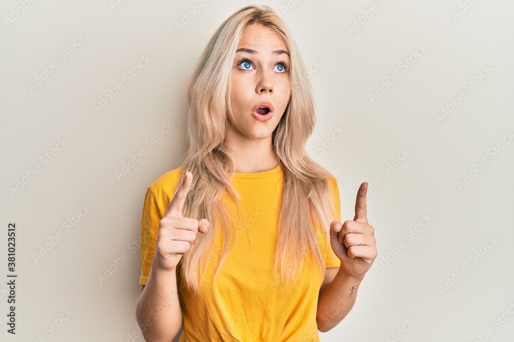 Beautiful caucasian blonde girl wearing casual tshirt amazed and surprised looking up and pointing with fingers and raised arms. Beautiful caucasian blonde girl wearing casual tshirt amazed and surprised looking up and pointing with fingers and raised arms.