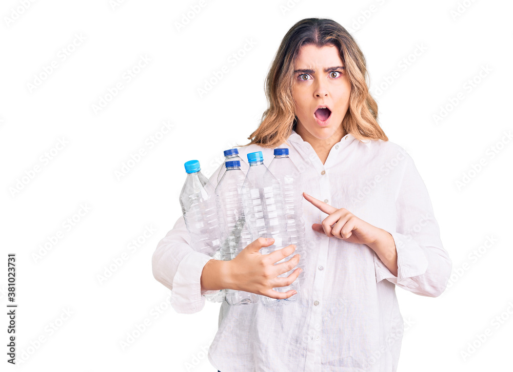 Young caucasian woman holding recycling plastic bottles scared and amazed with open mouth for surprise, disbelief face - obrazy, fototapety, plakaty 