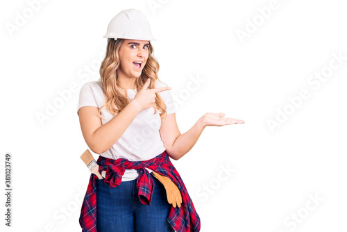 Young caucasian woman wearing security helmet amazed and smiling to the camera while presenting with hand and pointing with finger. photo