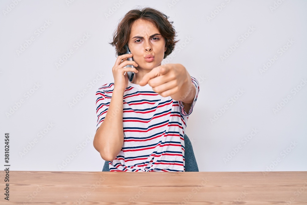 Young hispanic woman talking on the smartphone sitting on the table pointing with finger to the camera and to you, confident gesture looking serious