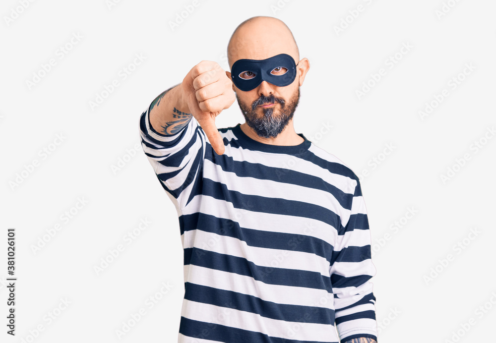 Young handsome man wearing burglar mask looking unhappy and angry showing rejection and negative with thumbs down gesture. bad expression. Young handsome man wearing burglar mask looking unhappy and angry showing rejection and negative with thumbs down gesture. bad expression.