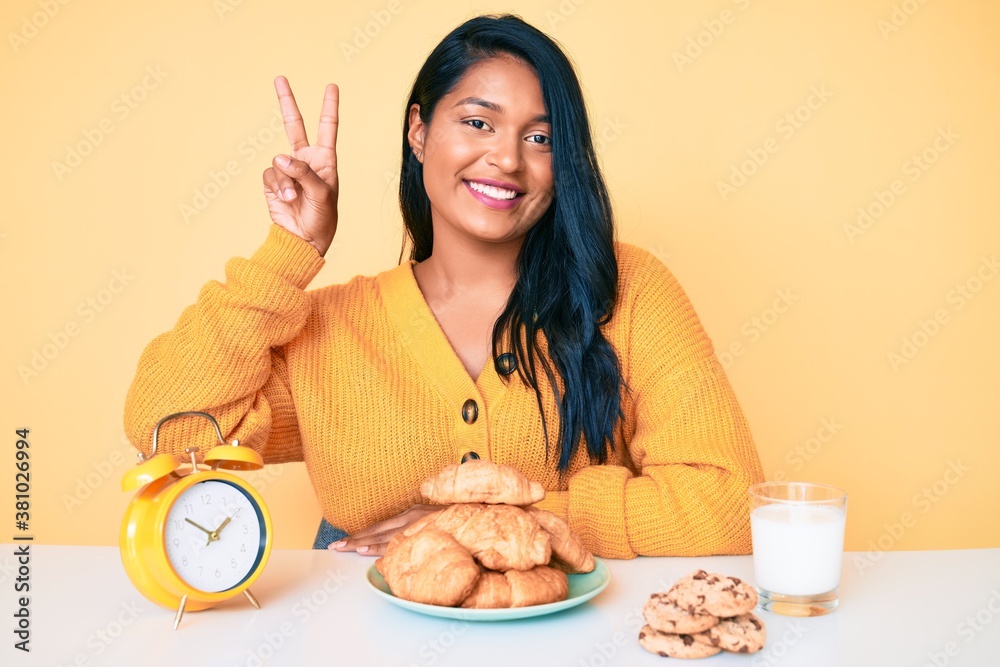 Beautiful latin young woman with long hair sitting on the table having breakfast smiling with happy face winking at the camera doing victory sign. number two.