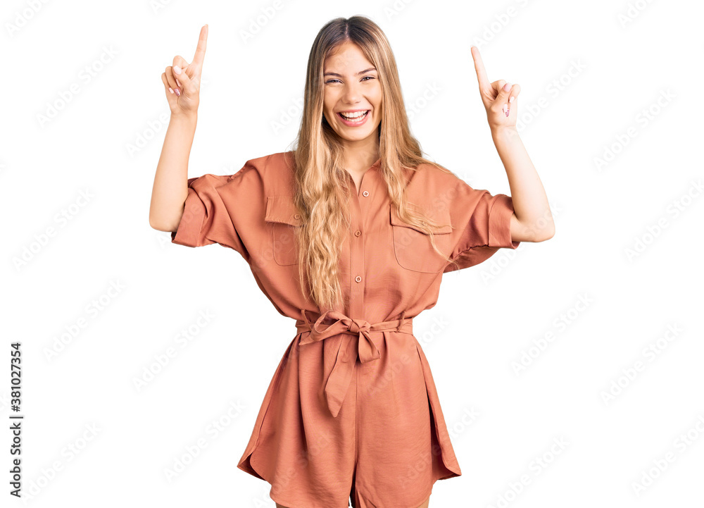 Beautiful caucasian woman with blonde hair wearing summer jumpsuit smiling amazed and surprised and pointing up with fingers and raised arms. Beautiful caucasian woman with blonde hair wearing summer jumpsuit smiling amazed and surprised and pointing up with fingers and raised arms.