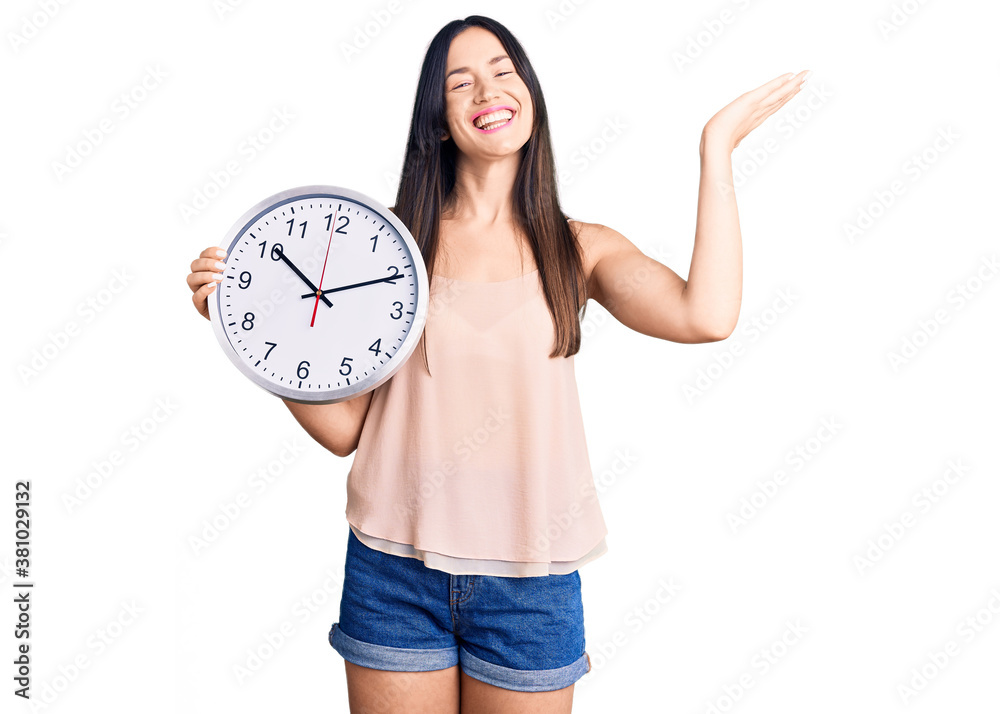 Young beautiful caucasian woman holding big clock celebrating victory with happy smile and winner expression with raised hands Young beautiful caucasian woman holding big clock celebrating victory with happy smile and winner expression with raised hands