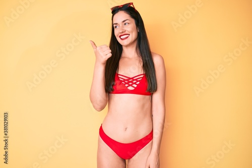 Young beautiful caucasian woman wearing bikini smiling with happy face looking and pointing to the side with thumb up.