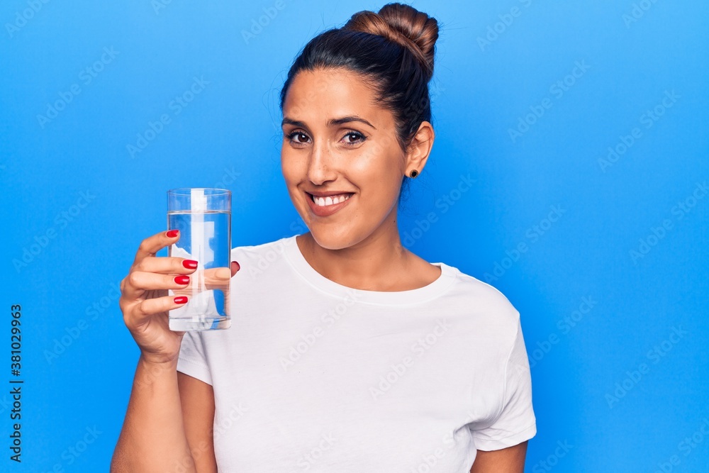 Young beautiful brunette woman drinking glass of water looking positive and happy standing and smiling with a confident smile showing teeth Young beautiful brunette woman drinking glass of water looking positive and happy standing and smiling with a confident smile showing teeth