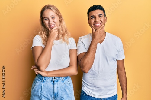 Young interracial couple wearing casual white tshirt looking confident at the camera smiling with crossed arms and hand raised on chin. thinking positive.