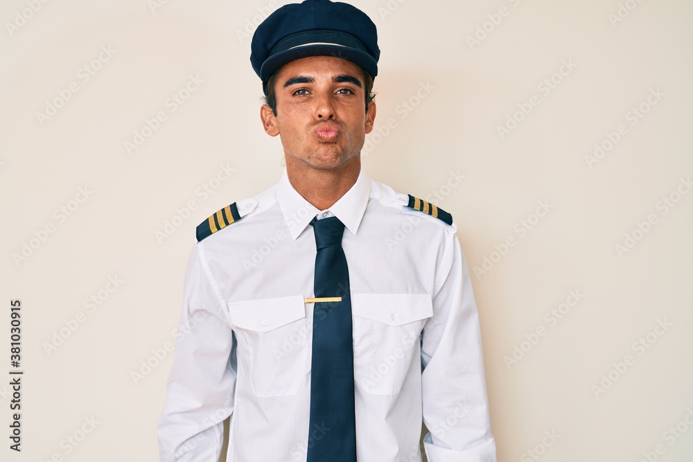 Young hispanic man wearing airplane pilot uniform looking at the camera blowing a kiss on air being lovely and sexy. love expression. Young hispanic man wearing airplane pilot uniform looking at the camera blowing a kiss on air being lovely and sexy. love expression.