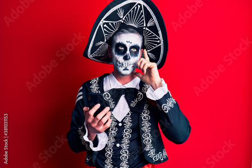 Young man wearing mexican day of the dead costume using smartphone serious face thinking about question with hand on chin, thoughtful about confusing idea