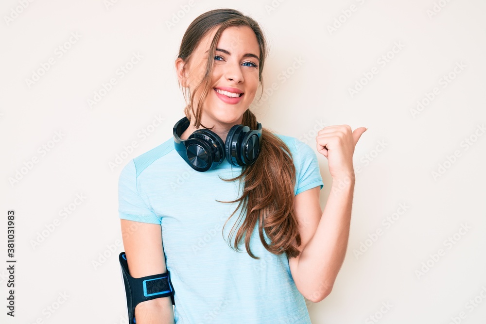 Beautiful young caucasian woman wearing gym clothes and using headphones smiling with happy face looking and pointing to the side with thumb up. Beautiful young caucasian woman wearing gym clothes and using headphones smiling with happy face looking and pointing to the side with thumb up.